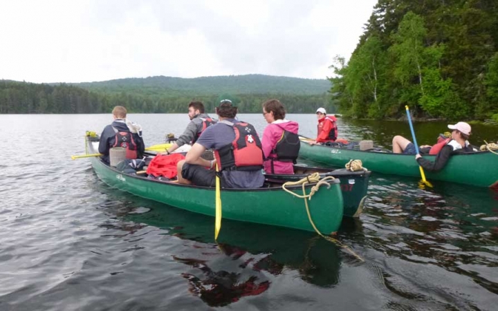 A group of young people wearing life jackets sit in canoes on calm water. The shores are covered in dense green trees.